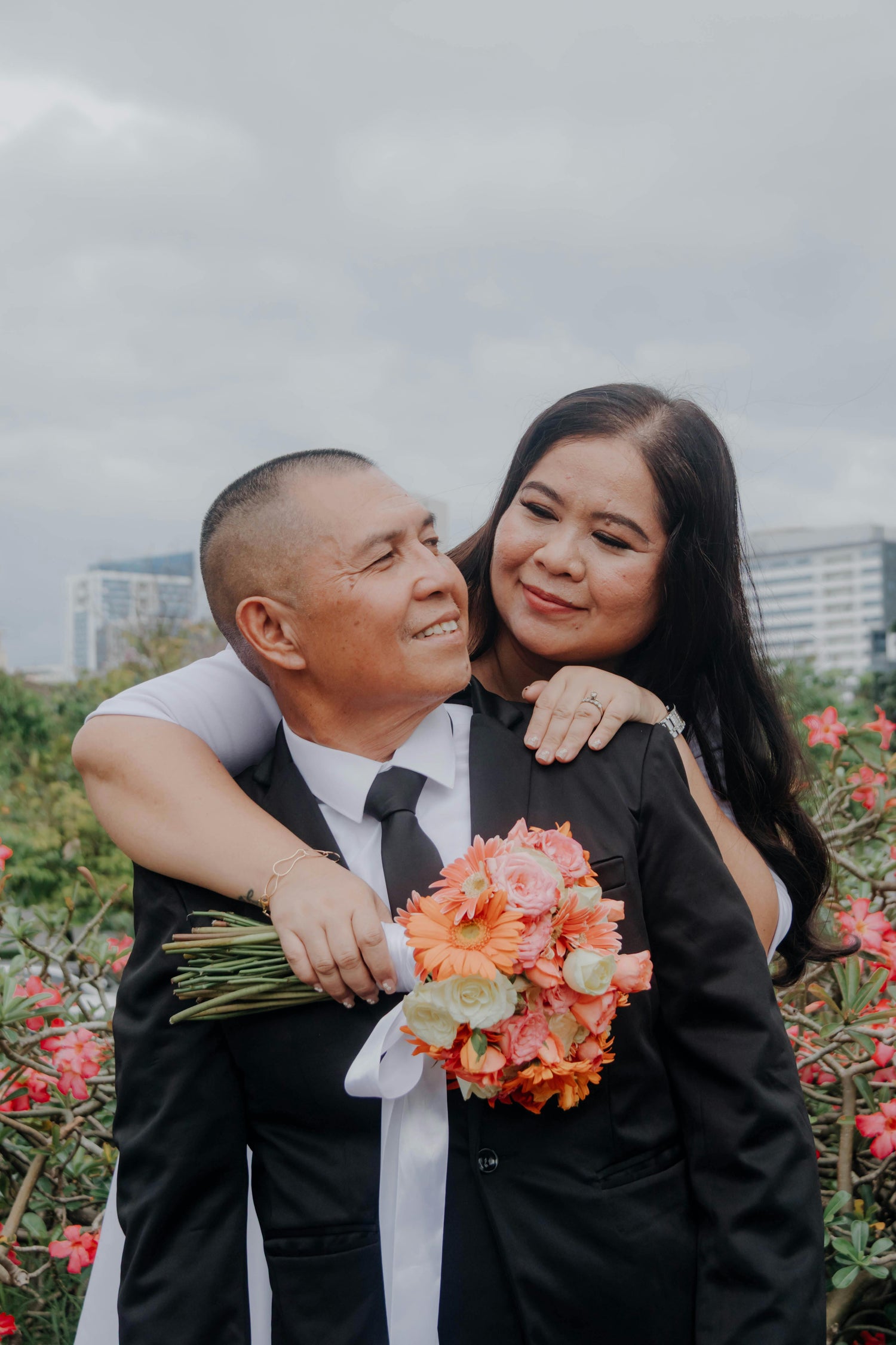 Man in a suit holding a woman with a bouquet of flowers, standing in front of a cityscape.