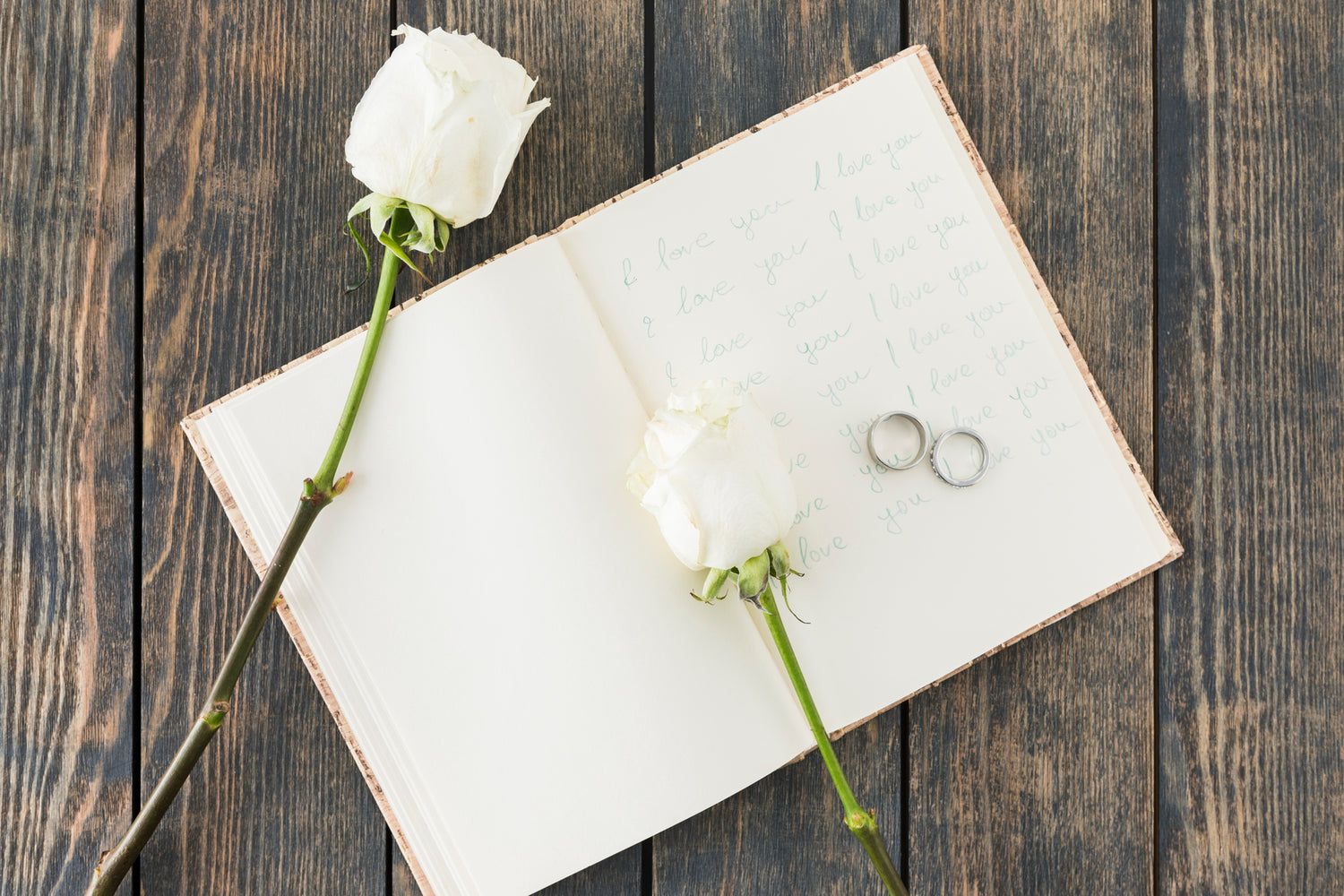 Open book with white roses and rings on a wooden surface